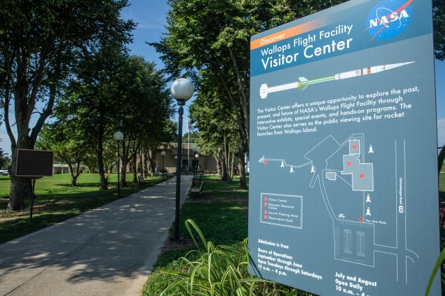 A gray sign with white text stands at the beginning of a tree lined sidewalk leading to a concrete building that houses the Wallops Flight Facility Visitor Center