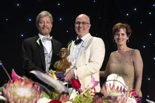 A group of people in formal wear poses with a small, copper-colored bust. The two men wear suits, one black and gray, one white and cream. The woman wears a gold dress. Colorful flowers cover the foreground.