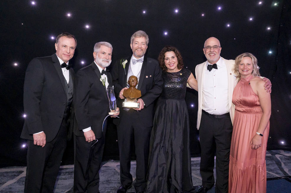 A group of people in formal wear stand on a stage, holding a small copper-colored bust and a crystal award. There are four men and two women. The backdrop is black with sparkling stars.