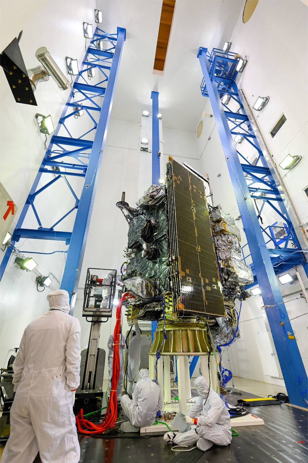 The GOES-U satellite stands vertical in a clean room on a stand made up of many electronic components and covered in static bags, its solar panels facing out. Many technicians in white clean suits work around the bottom of the satellite.