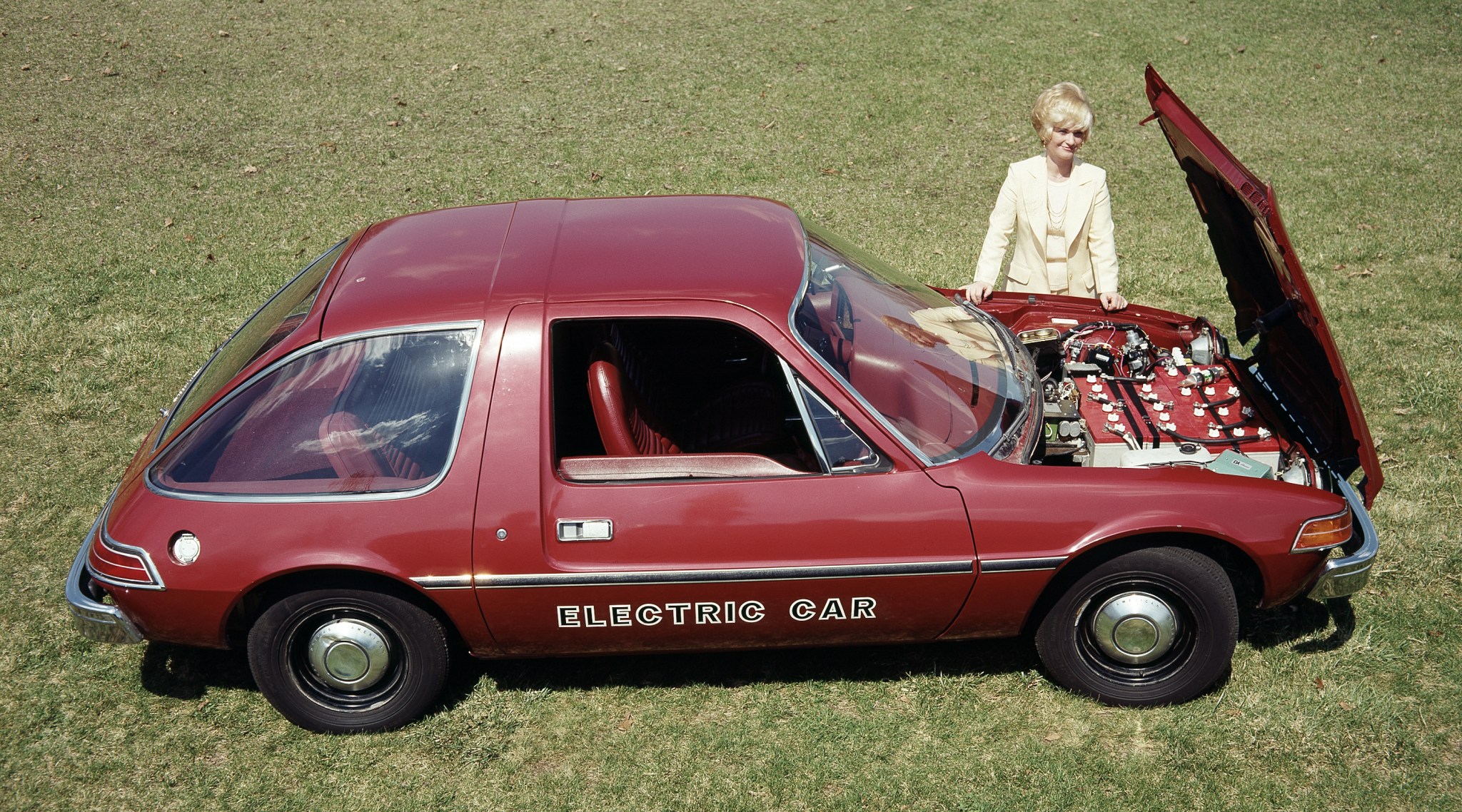 Woman standing next to car in grass field with hood open.