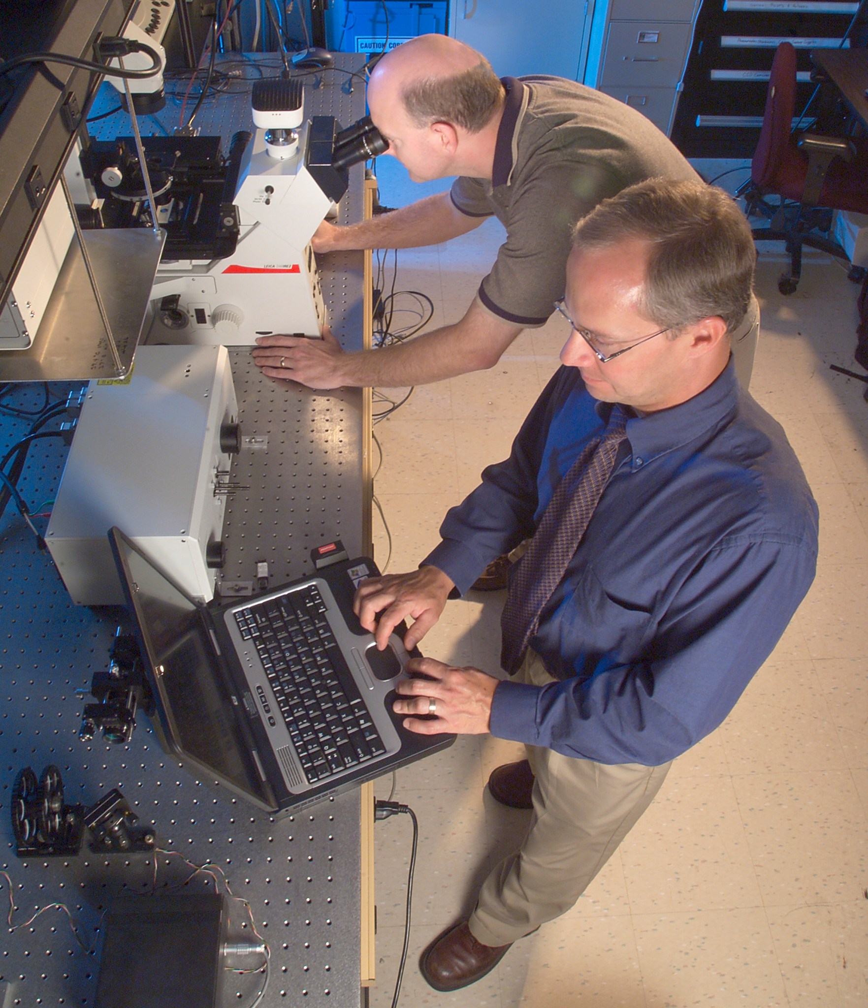 Two men in lab working on laptop and microscope.