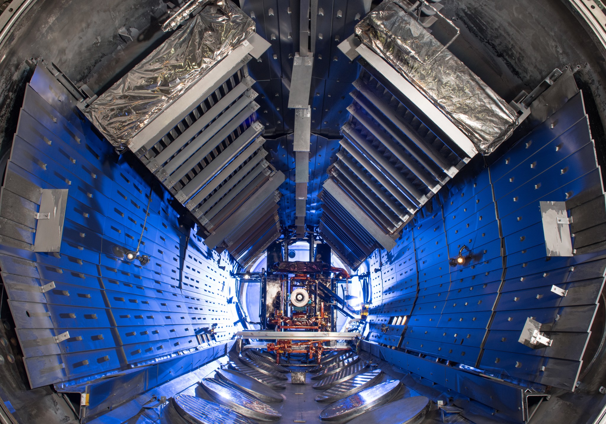A view inside a space vacuum chamber. The interior of the chamber resembles a large metal cylinder with silver, rectangular, panels lining the walls and ceiling. The interior of the chamber is illuminated by blue lights. Test hardware is mounted in the center of the chamber on a structure that resembles scaffolding. 