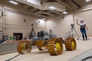 A test version of the VIPER rover is seen inside a large bin of lunar simulant. The bin and its contents resemble a large sandbox. A NASA engineer stands inside the bin with an arm stretch above her head. She wears a white protective respiratory hood.