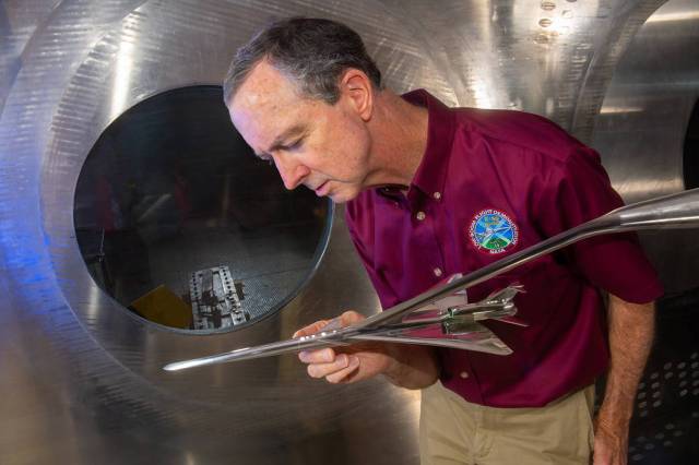 A man in a red shirt leans forward to look closely at a metal airplane model installed in a wind tunnel.