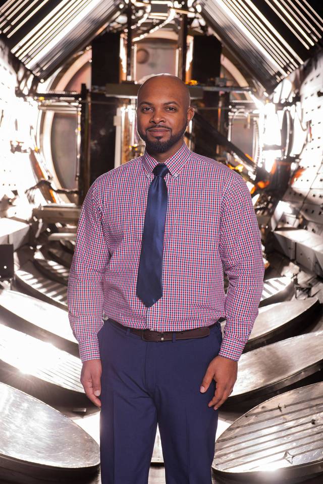 Man in shirt and tie looks at camera while standing inside a laboratory vacuum chamber.