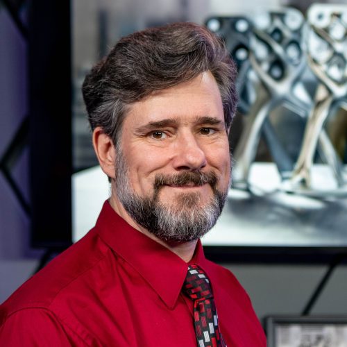 man in red short with beard in front of display of shiny metal parts