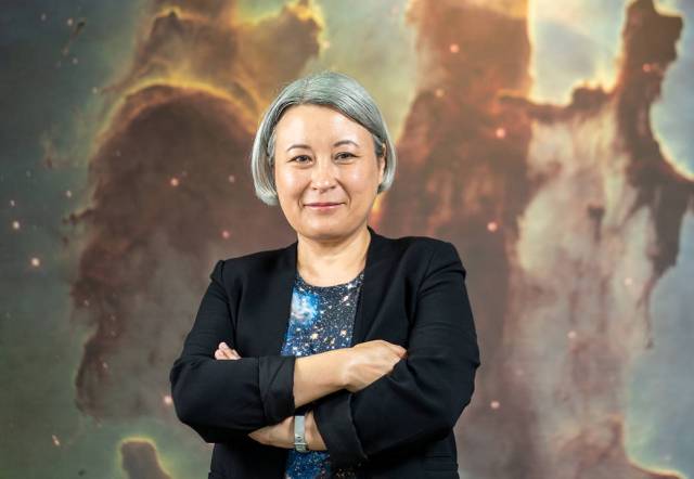 A woman with a short grey bob softly smiles with crossed arms. She's dressed in a starry blue shirt and black blazer& stands in front of the Pillars of Creation image. Tentacles of dark space dust and gas climb upwards, with starlight dotting the image. 