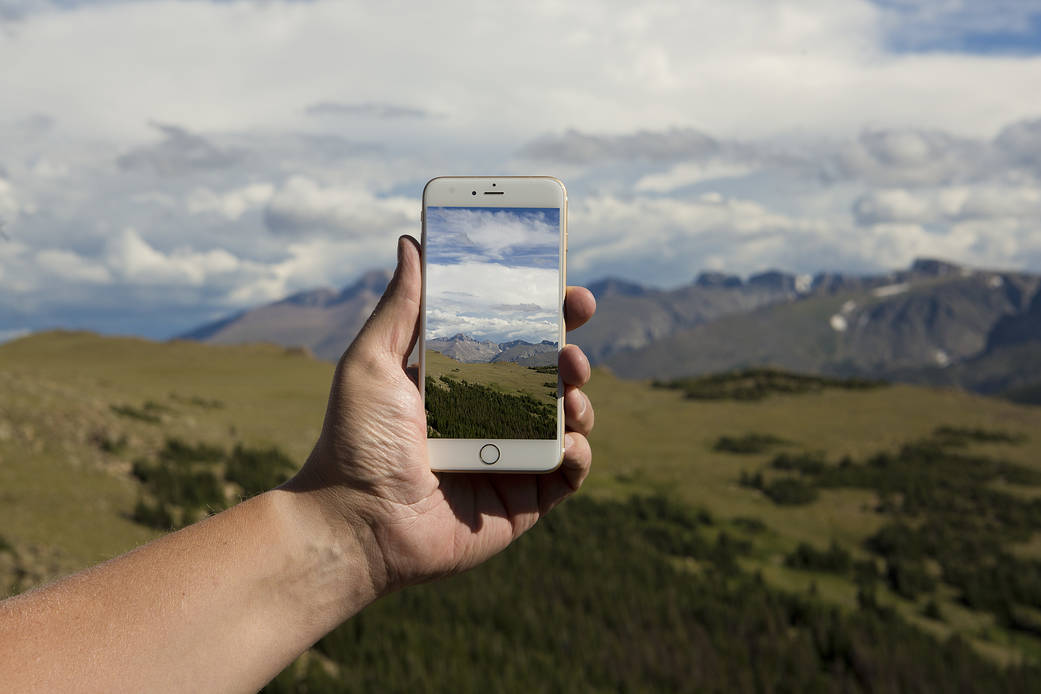 A person's hand holds a cellphone using the globe observer app and is taking an image of a grey mountain range far away. Green rolling hills line the foreground of the image and the person's hand extends from the left-hand side. 