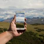 A person's hand holds a cellphone using the globe observer app and is taking an image of a grey mountain range far away. Green rolling hills line the foreground of the image and the person's hand extends from the left-hand side. 