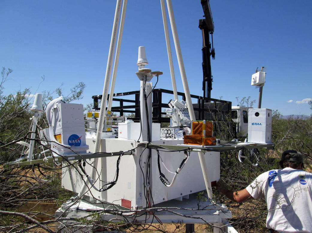 Outdoors in a brush-filled area, a man stands next to a large white box with student experiments attached.