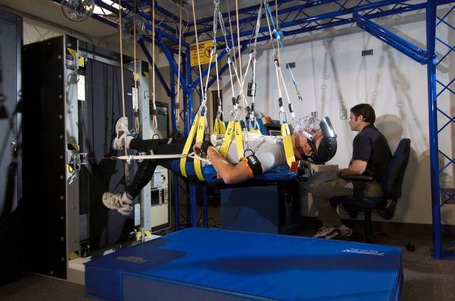A male test subject jogs on a vertical treadmill while held in the air by harnesses