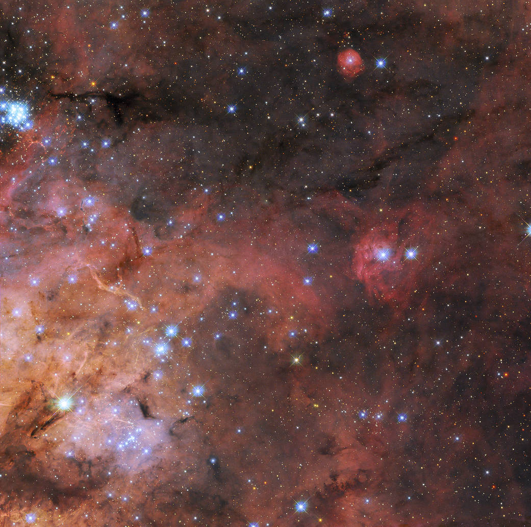 Wispy, nebulous red clouds extend from lower left. At top and right: dark background of space is seen through sparse nebula. Along the left, layers of brightly colored gas and dark, obscuring dust, and a cluster of small, bright blue stars at upper left.