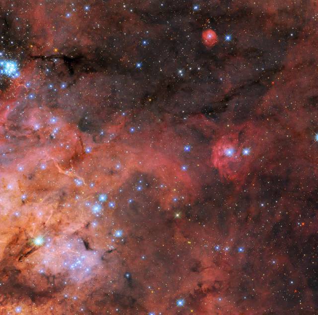 Wispy, nebulous red clouds extend from lower left. At top and right: dark background of space is seen through sparse nebula. Along the left, layers of brightly colored gas and dark, obscuring dust, and a cluster of small, bright blue stars at upper left.