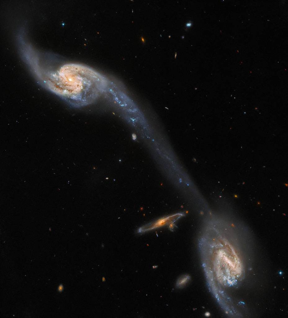 Two face-on spiral galaxies at upper-left and lower-right corners. A long, faint, pale-blue streak joins them, crossing the field diagonally. A small, orange, edge-on spiral galaxy at left of the lower galaxy.