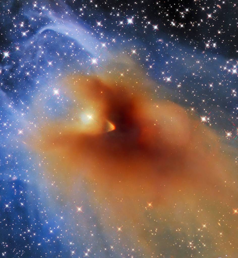 An irregularly shaped bright orange cloud dense gas and dust, which appears darker and more compact at center and is outlined by thinner gas and dust in light shades of blue. A multitude of bright stars against a black background.