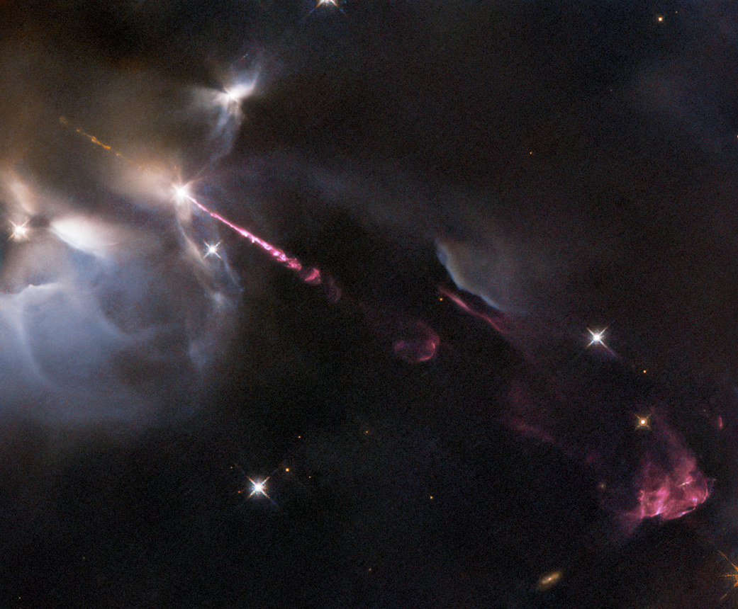 Dark cloud of gas and dust a with bright-white glowing outline in the upper left. Pinkish-red jet of material extends from the cloud to the lower right where billowing reddish-pink cloud forms. background stars