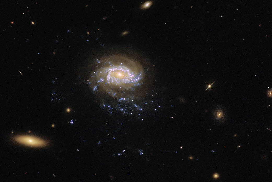 Center left: spiral galaxy with large, faint, reddish arms, and a bright, reddish core over two brighter blue spiral arms. They hold patches of star formation that form long trails down, resembling tendrils. Larger elliptical galaxy in lower-left corner.