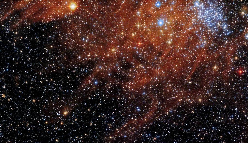 Bright blue-white cluster of stars at upper right, surrounded by field of dimmer stars and rusty-red gas and dust clouds