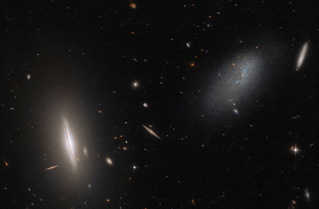 Right of center: faint, scattered collection of cool stars, an irregular galaxy. Lower left: disk-shaped galaxy nearly edge-on lies surrounded by a wide glow. Smaller galaxies in various orientations cluster around the two. Black background.