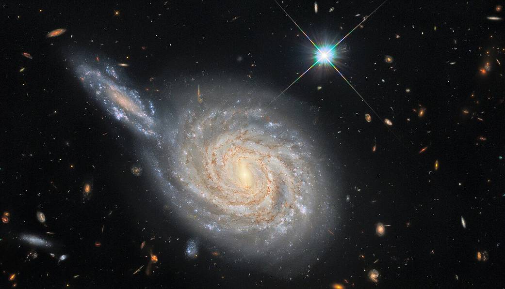 Spiral galaxy at center, with an edge-on galaxy at upper left (10 o'clock) that appears to be crashing into the spiral. Bright-blue star to the upper right. Many fainter galaxies in the background