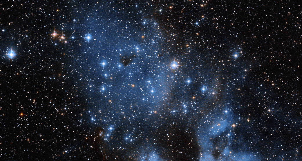 Bright, blue-white stars of varying sizes fil the scene. A smattering of orange-white stars dotted throughout. A swath of pale-blue gas and dust extends from upper left to lower right across the image.