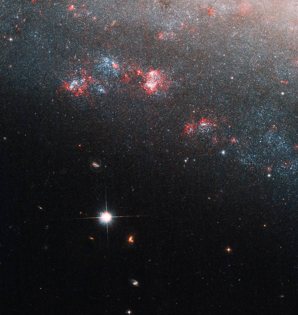 a portion of the galaxy fills the upper right half of the image, blue and red clusters of stars and gas dot its edge, a bright foreground star sits to the lower left of the galaxy