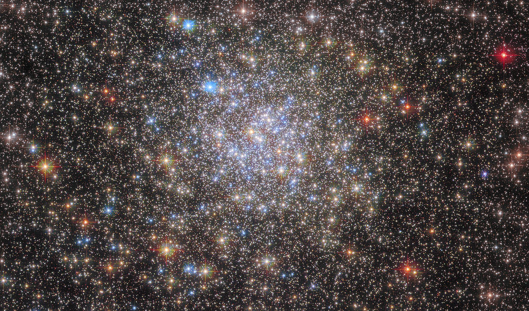 Stars fill the view. A dense, spherical collection of blue and yellow-white stars toward the center. The image’s edges hold redder foreground stars, and many small background stars.