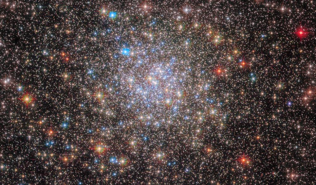 Stars fill the view. A dense, spherical collection of blue and yellow-white stars toward the center. The image’s edges hold redder foreground stars, and many small background stars.