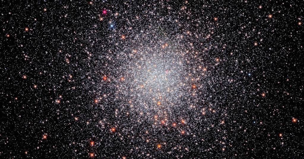 Center of the image is filled with a sphere of white stars. A smattering of orang-white stars dots the sphere. They are set against a black background that holds a smattering of stars.