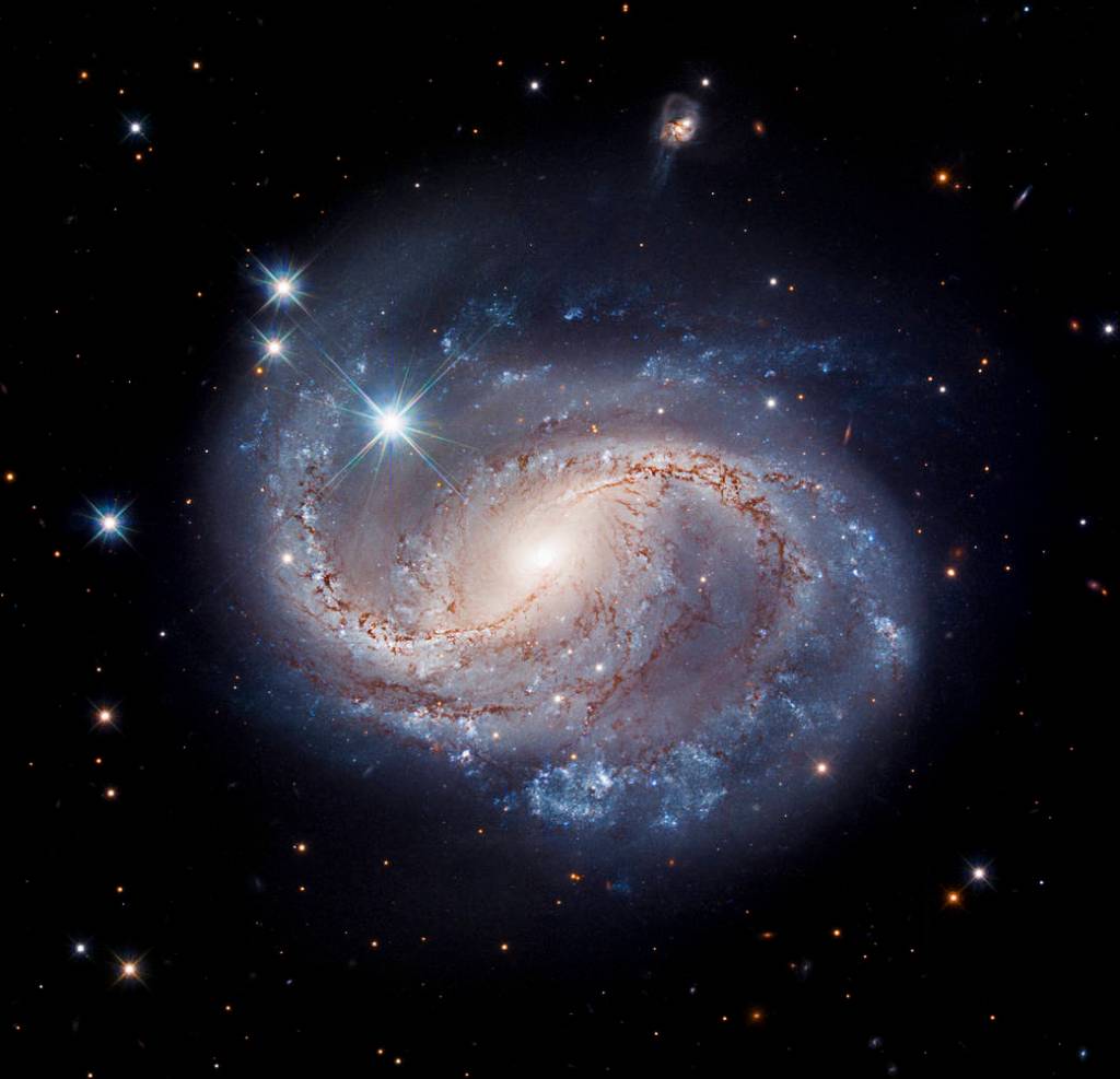 Image center: blue, and pinkish-white swirls of the barred spiral galaxy NGC 6956. Dark, reddish-brown dust lanes along the inner part of the spiral arms. Inky black background with foreground and distant stars and galaxies.