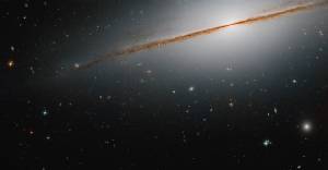 Edge-on galaxy, with a distinctive dust lane, extending from upper right to middle-left of the image. Distant galaxies dot the scene.