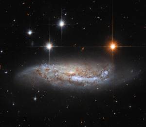 barred spiral galaxy seen from the side. 3 bright-white foreground stars are to the upper left of the galaxy, one bright-orange-white star is to the upper right.