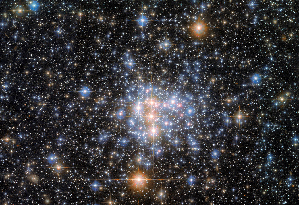 Bright stars cluster at image center. They shine in shades of white, blue, and orange, and have diffraction spikes. The stars grow more sporadic at the image’s edges, and several smaller, more distant stars glow against a black backdrop of space.