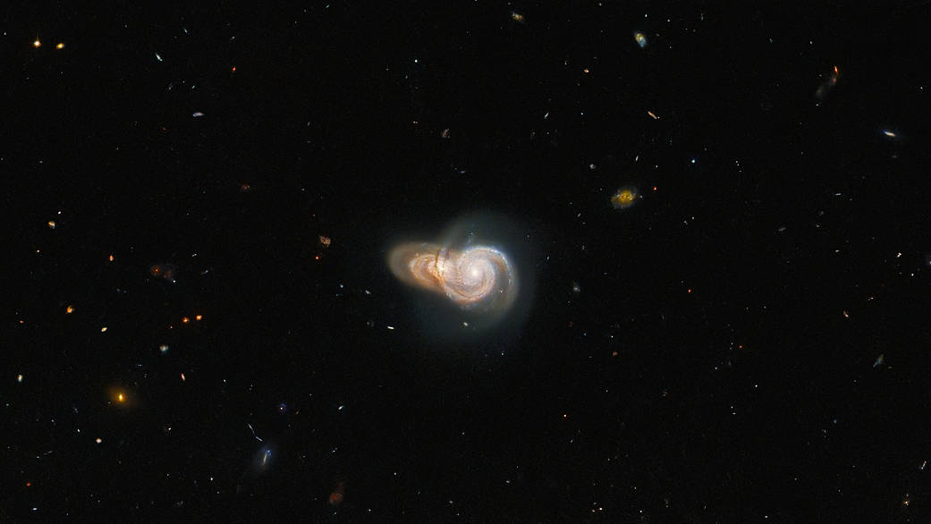 center of image holds two spiral galaxies. The foreground galaxy is face-on, while the one behind it is at a slight angle. Both appear yellow-gold with streams of bright white stars.