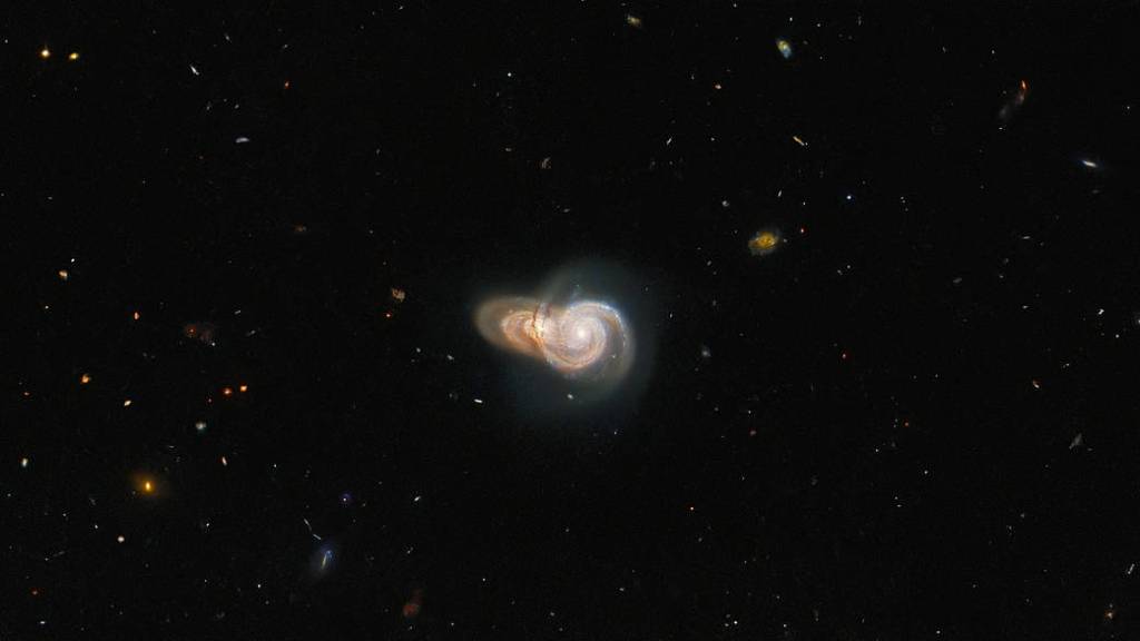 center of image holds two spiral galaxies. The foreground galaxy is face-on, while the one behind it is at a slight angle. Both appear yellow-gold with streams of bright white stars. 