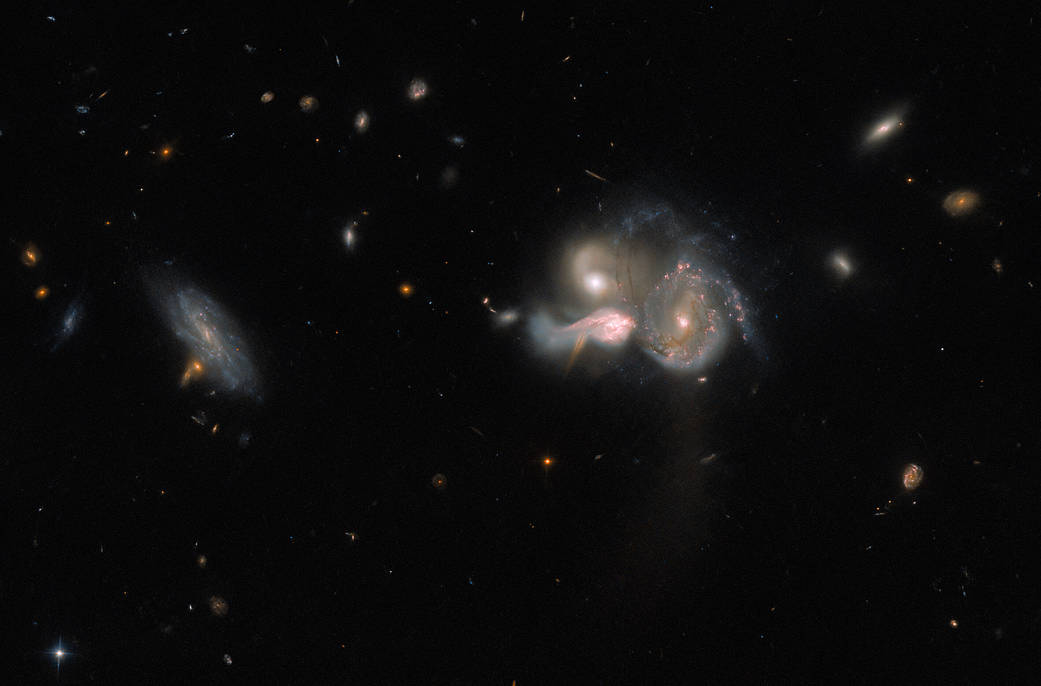 Just right of center: 3 galaxies appear to be merging. Their shapes are distorted, with strands of gas and dust running between them. Far-left: an unconnected, dimmer spiral galaxy. Dark background with a few smaller, faint galaxies and a couple of stars.
