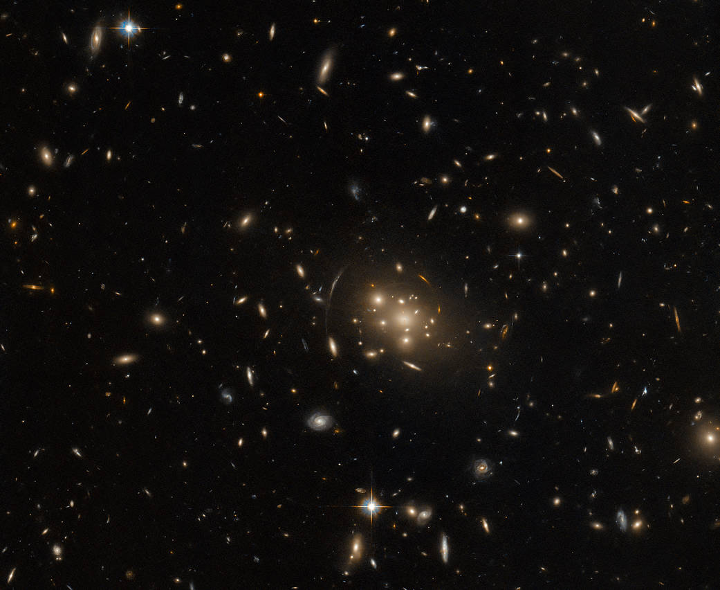 A cluster of large galaxies, surrounded by stars and smaller galaxies on a dark background. The central cluster is mostly made of bright elliptical galaxies. Near the cluster is the stretched, distorted arc of a gravitationally lensed galaxy.