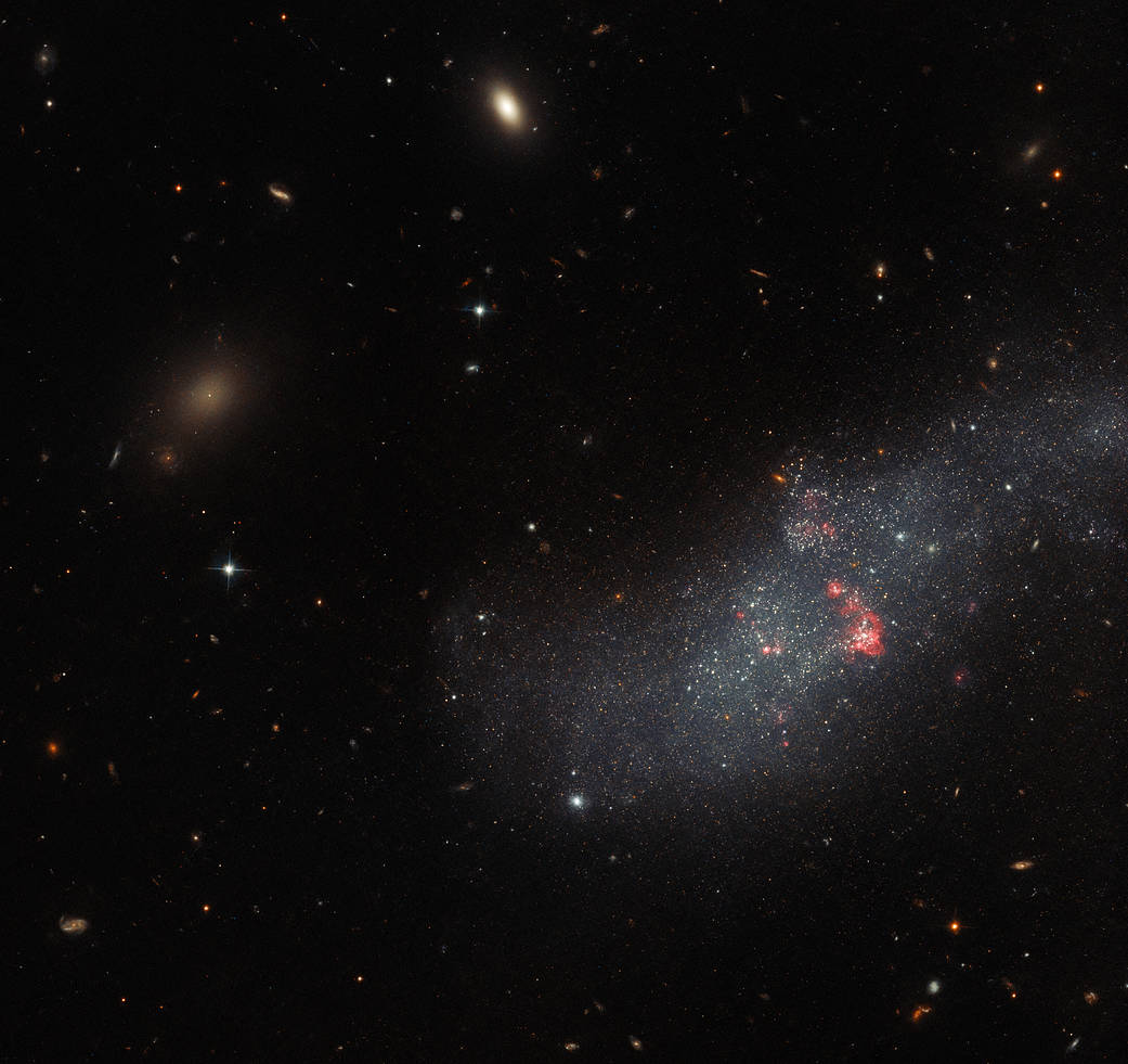 Band of bluish light speckled with stars and a few small red clouds of gas extends from image center to the right side. Background is black, with small galaxies and stars. 
