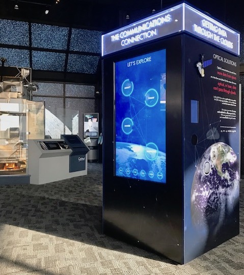 A photo of a museum exhibit, showing a tall black box with screens displaying information about space communications and navigation, with photos of Earth and text on the sides. A wall with glowing images of numerous stars is in the far background, with other exhibits partly visible.