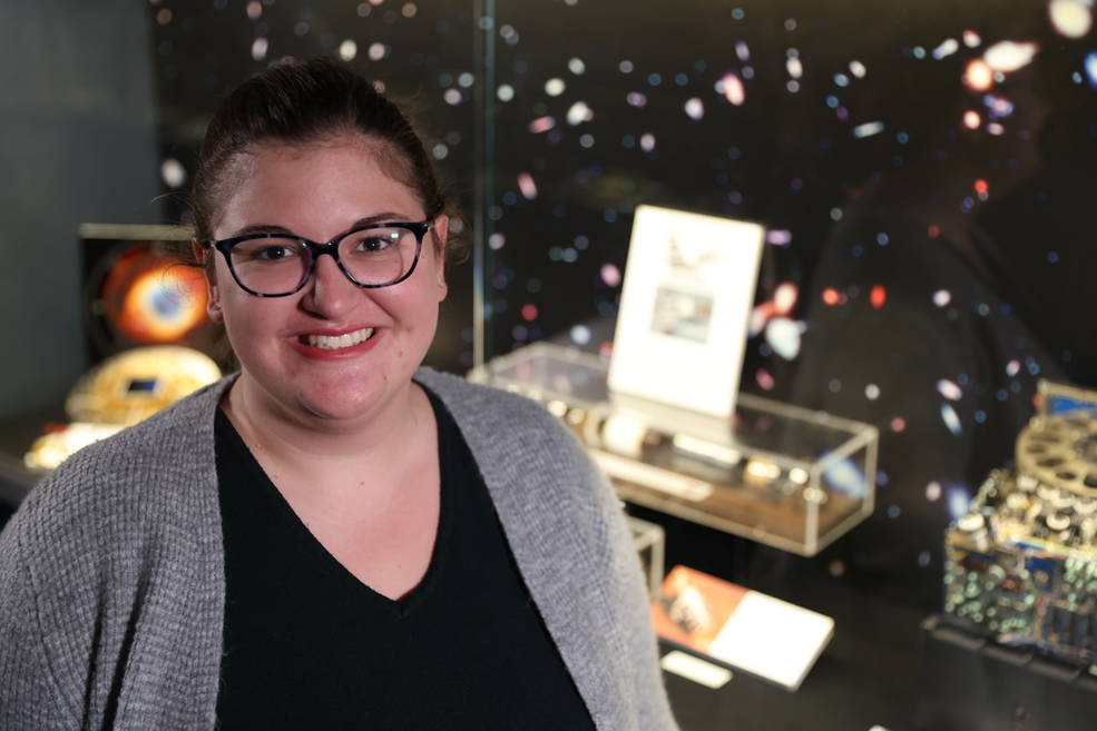 Hannah Daelman, a woman with dark hair pulled back, smiles in a portrait. She wears a black tee, gray sweater, and glasses. An exhibit and black, galaxy-sprinkled backdrop are visible in the background.