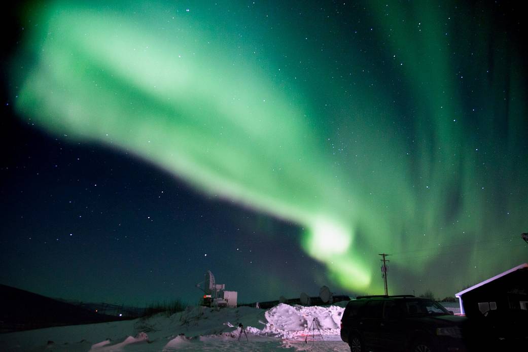 The night sky is filled with the green northern lights above a small snowy building.