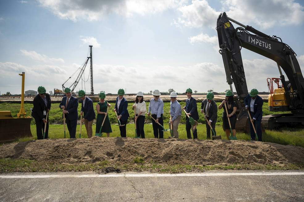 A group of men and women stand in front of an empty field with green and white hard hats and shovels. An excavator sits off to the side.