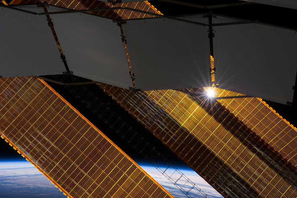 The sun shines through a truss-based radiator panel and a primary solar array panel on the Earth-orbiting International Space St