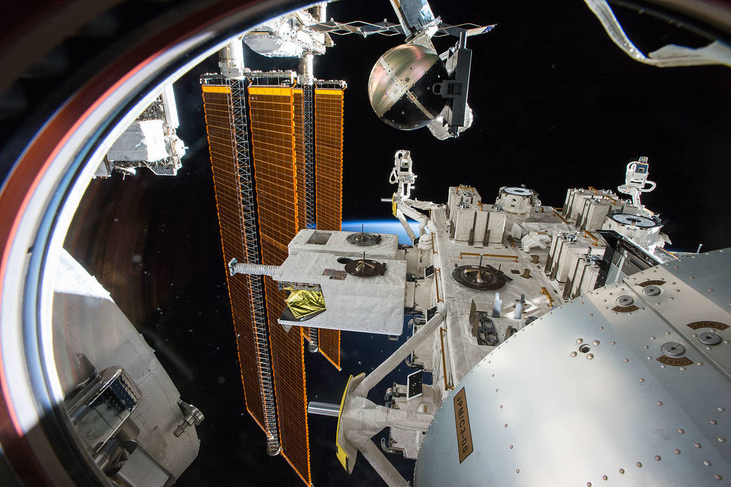 View from space station window showing solar arrays at left and round satellite on deployer