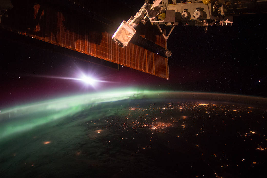 Aurora in early morning on Earth's horizon with city lights below and space station solar arrays above