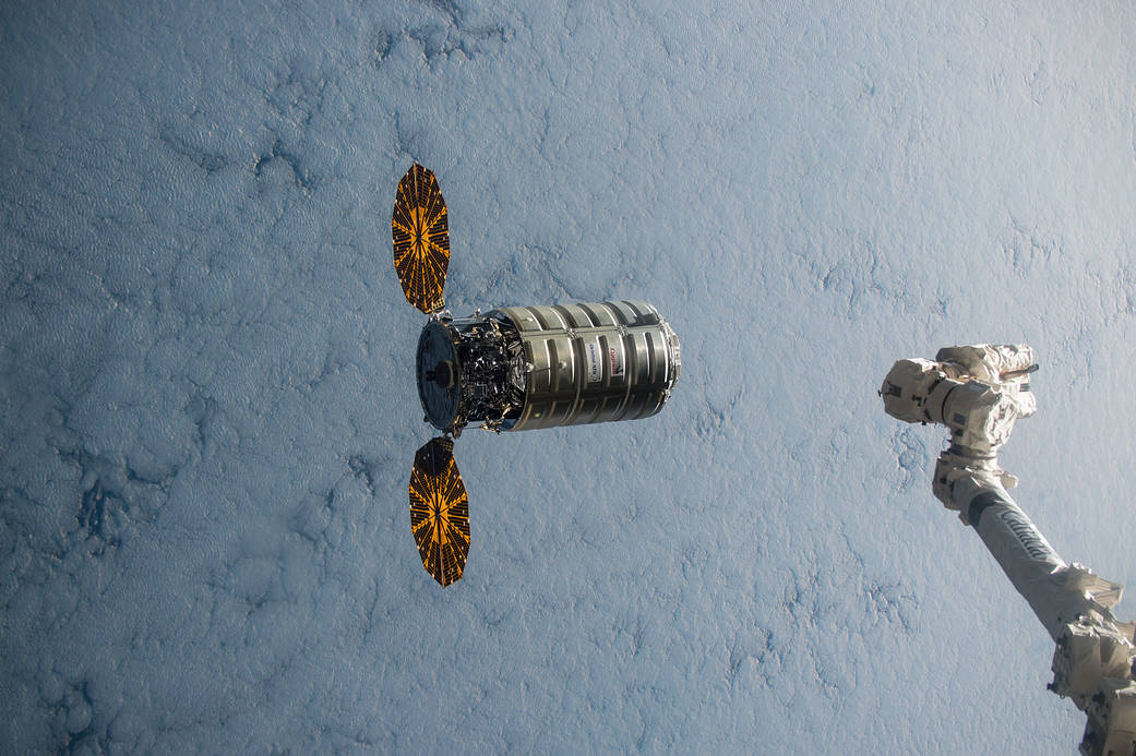 Cygnus spacecraft with solar arrays deployed and clouds below approaching robotic arm in lower right