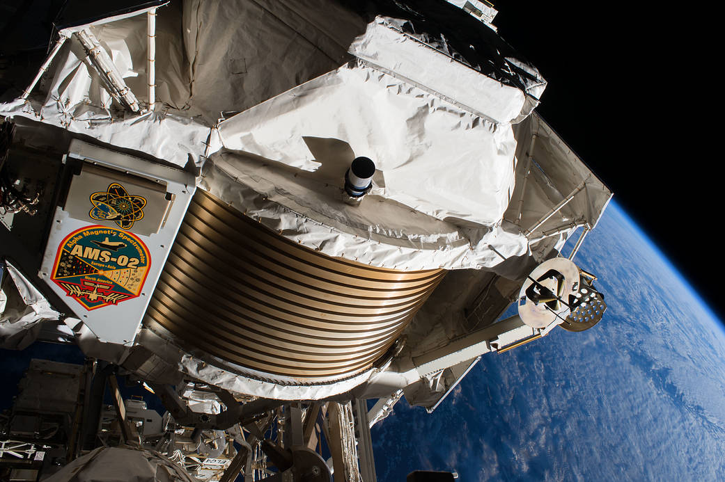 A series of gold bands is visible on the front of the AMS, with white material covering the top, an arm with a rotor at the end to the right, and a panel to the left bearing the investigation logo. Other equipment is visible below AMS, with a cloudy Earth in the background.