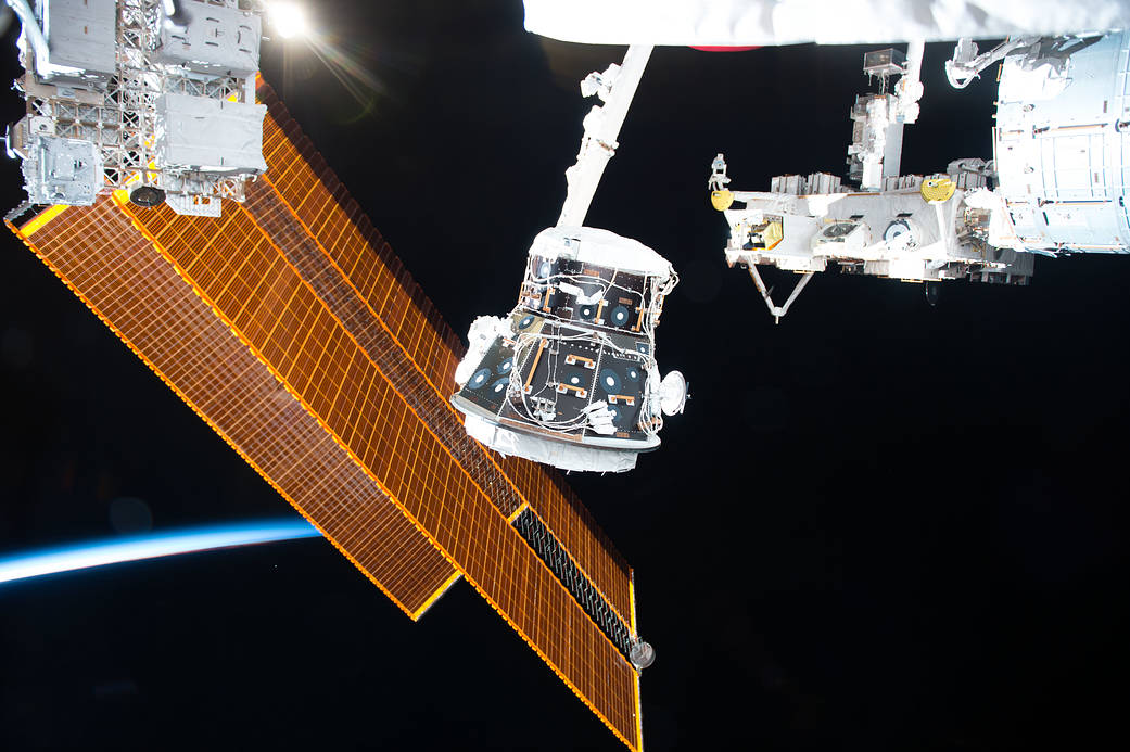 Adapter held by robotic arm outside space station with solar array visible at left