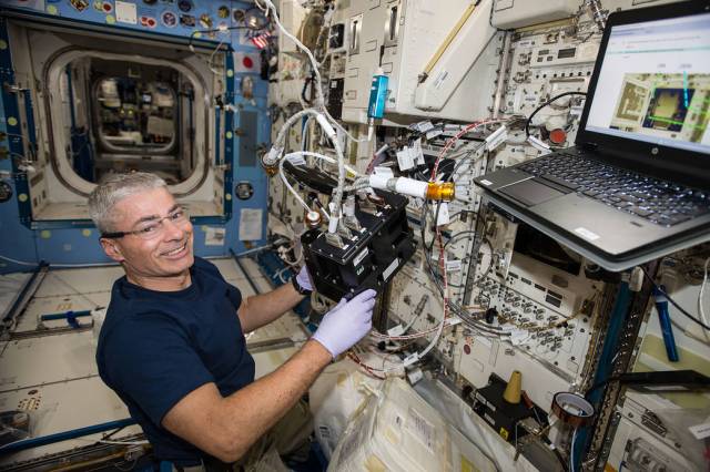 Mark Vande Hei looks happy, smiling at the camera in a navy blue t-shirt, purple surgical gloves, and metal-rimmed glasses. Aboard the ISS, he holds the Advanced Nano Step Cartridge in the Solution Crystallization Observation Facility during installation.